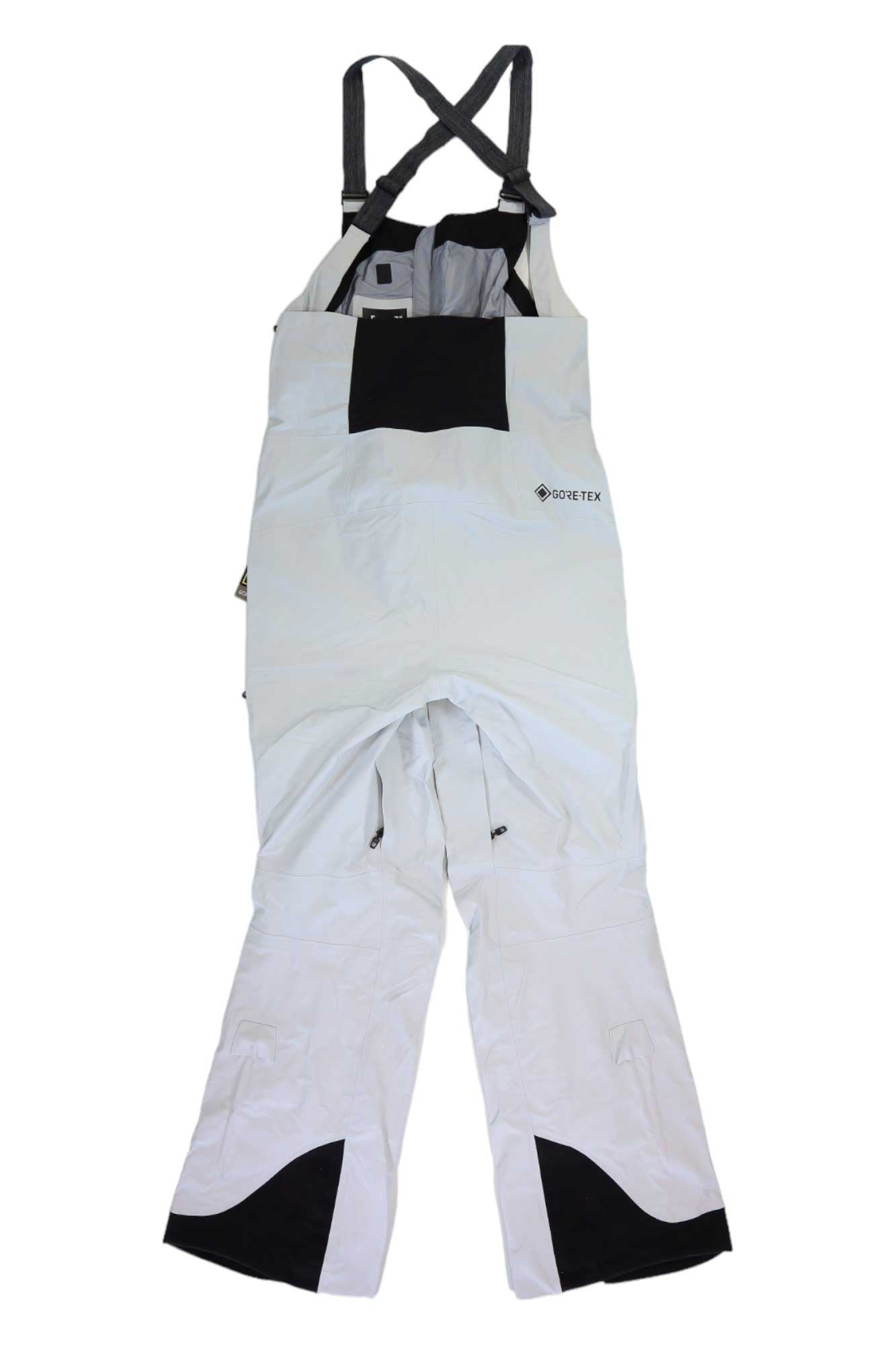 White waterproof Burton Womens [ak] Gore-Tex Kimmy Bib Pant with black shoulder straps, front chest pocket, and reinforced ankle patches, laid flat with legs extended and straps crossed at the top.