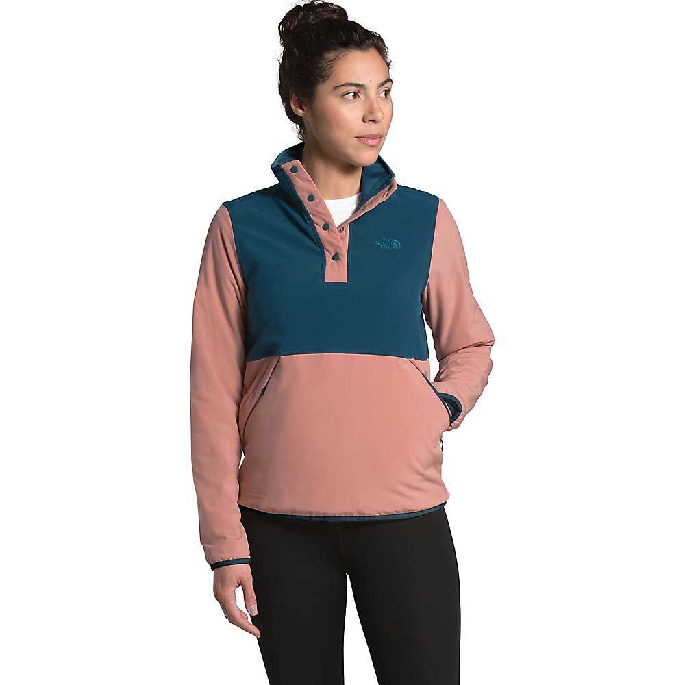 A woman wears the North Face Womens Mountain Sweatshirt Pullover 3.0 with a teal top and dusty pink bottom, paired with black leggings. She stands with one hand in her pocket, hair tied up, looking to the side.