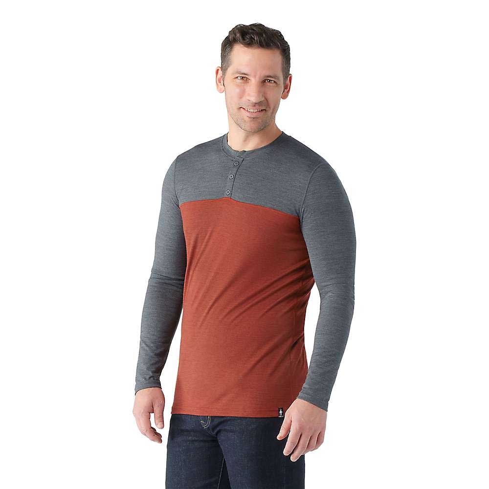 A man models the Smartwool Men's Merino Sport 150 Henley, featuring a moisture-wicking design with grey sleeves and upper section, rust-colored lower section, and stands smiling against a plain white background.