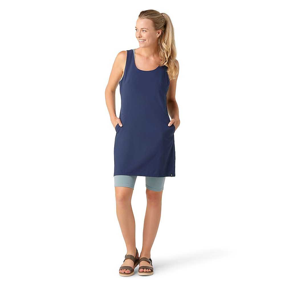 A woman smiles in the Smartwool Womens Intraknit Active Dress over light blue bike shorts and brown sandals. Her hair is in a ponytail, and her hands are in her dress pockets against a plain white background.
