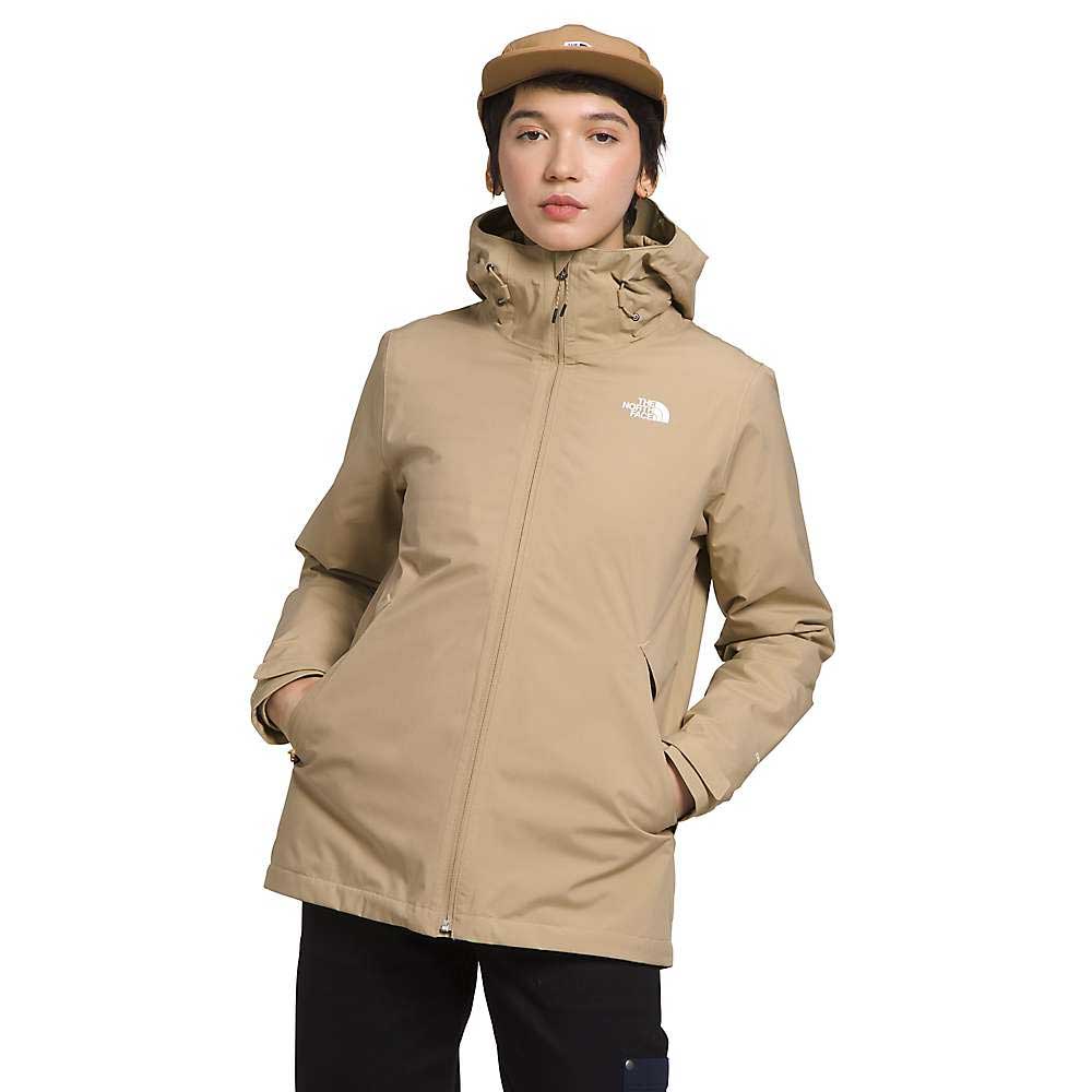 A person in The North Face Women's Carto Triclimate Jacket and matching tan cap stands with hands in pockets against a plain white background. The tan jacket, featuring the logo on the chest, is made from recycled polyester.