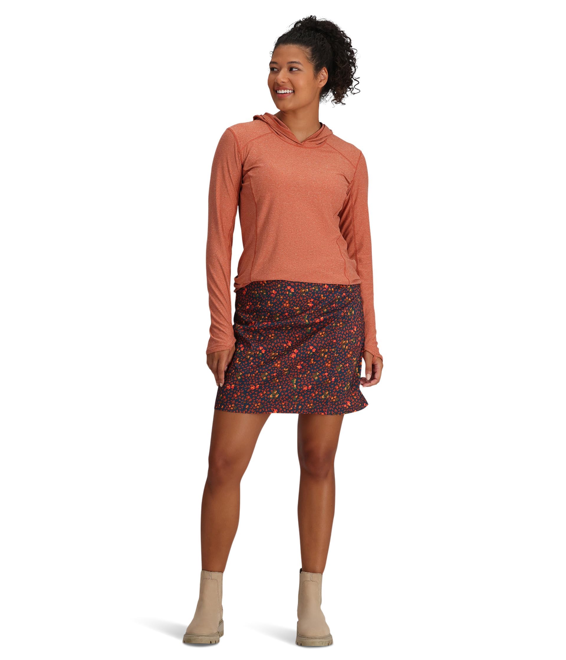 A woman smiles, wearing a long-sleeved rust hooded top, beige ankle boots, and a Royal Robbins Women's Discovery III Printed Skort in Navy (Size 8), which offers UPF 40 sun protection, against a white background.