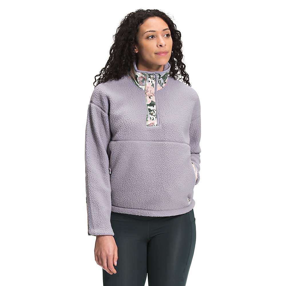 A woman with curly hair wears The North Face Womens Cragmont Fleece 1/4 Snap Top in light purple with a floral-patterned collar and dark green pants, standing against a white background.