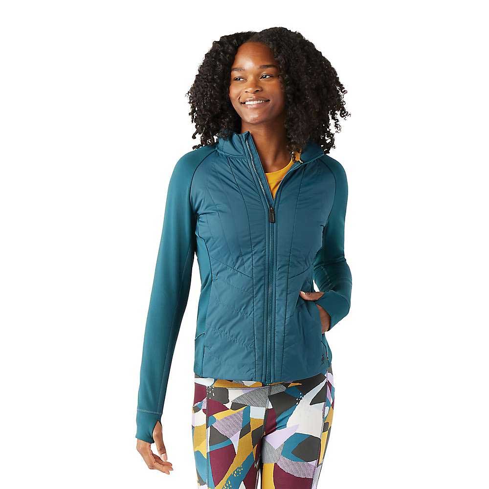 A woman with curly hair smiles while wearing a teal Smartwool Womens Smartloft Jacket with Merino wool insulation and colorful patterned leggings against a white background.