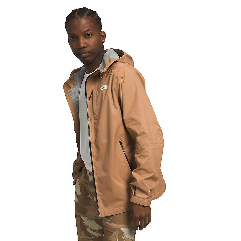 A man wears The North Face Men's Alta Vista Jacket, a tan hooded waterproof jacket made from recycled fabrics, along with camouflage pants, looking toward the camera against a plain white background.