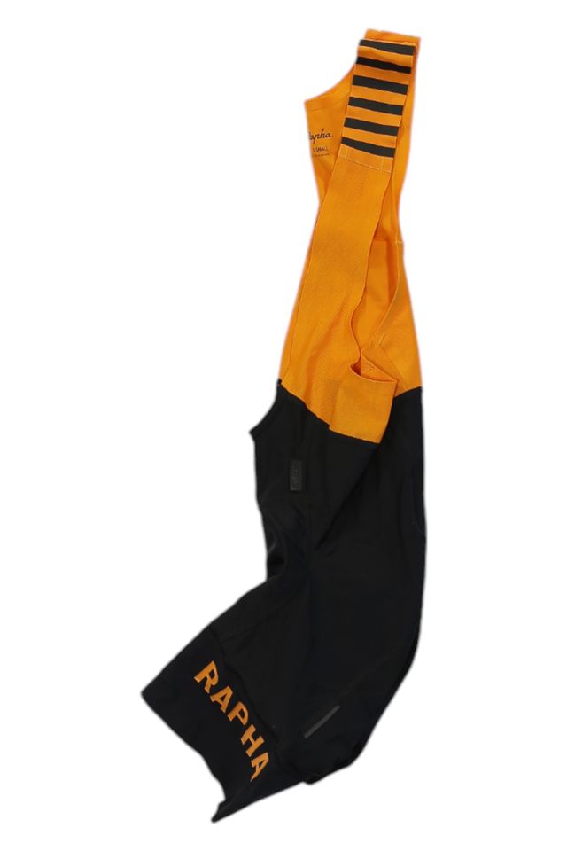 A pair of Rapha Mens Pro Team Bib Shorts II - Regular, with orange straps featuring black horizontal stripes and a black lower half. "RAPHA" is printed in orange on one leg. Race-grade chamois. Set against a white background.