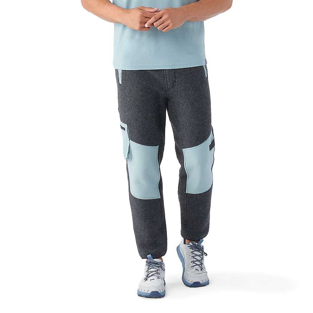 A person wearing Smartwool Men's Hudson Trail Fleece Pant in gray with light blue knee patches, a matching light blue shirt, and white athletic shoes stands with hands clasped. The photo shows the outfit from shoulders to feet.
