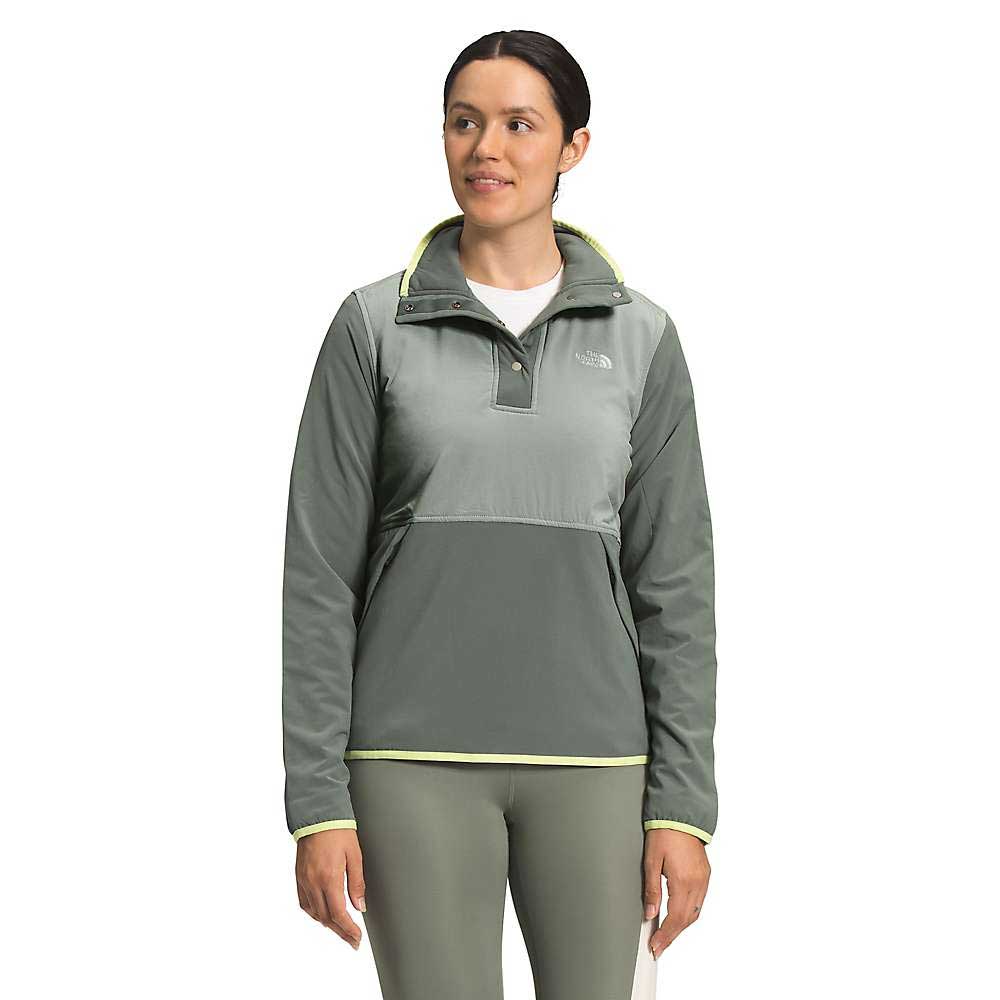 A woman stands against a plain white background, wearing the North Face Womens Mountain Sweatshirt Pullover 3.0 in two-tone green with a high collar and yellow trim, made from recycled polyester, looking slightly to the side.