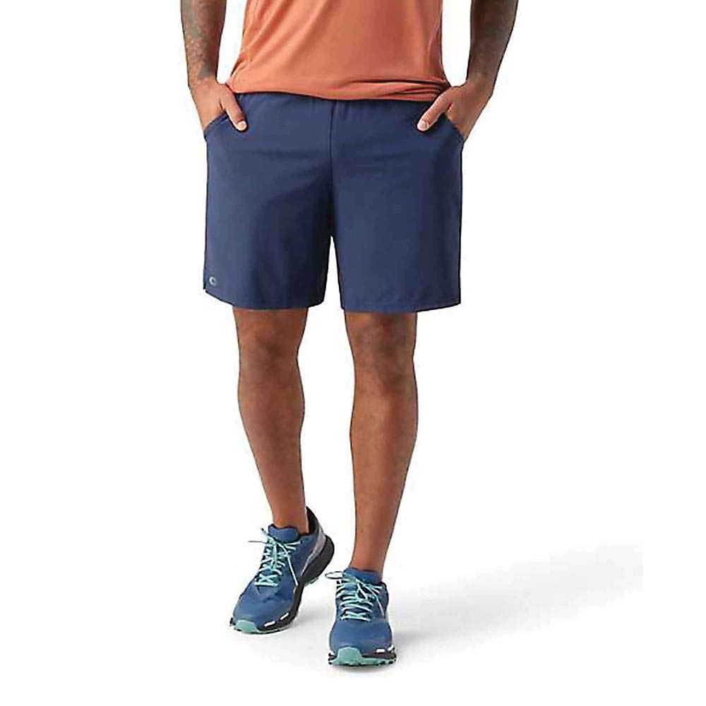 Wearing Smartwool Men's Active Lined 8 Inch Shorts in navy, a salmon t-shirt, and teal running shoes, a person stands with hands in their pockets against a plain white background. Their upper body is not visible.