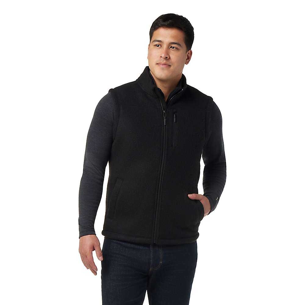 A man, wearing the Smartwool Men's Hudson Trail Fleece Vest over a dark long-sleeve shirt and jeans, stands against a white background with one hand in his pocket, looking slightly to the side.