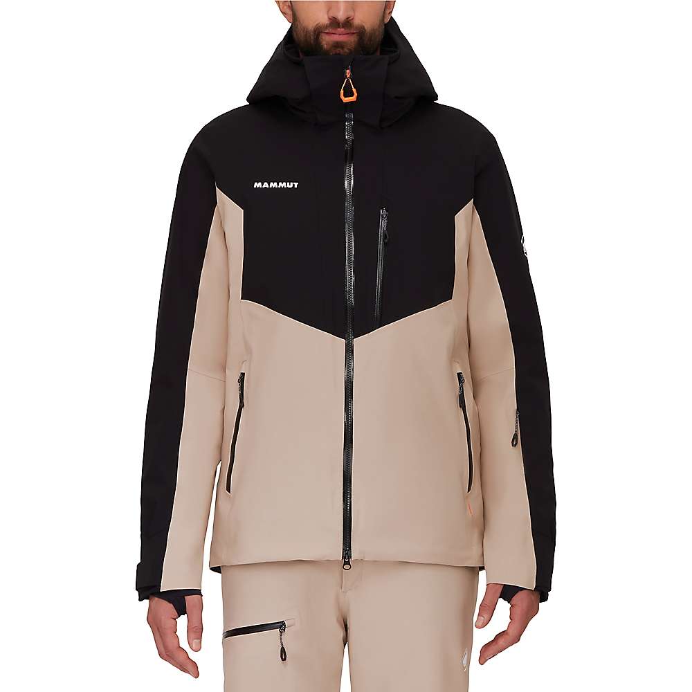 A person wearing a beige and black Mammut Mens Stoney HS Thermo Jacket, featuring a hood, front zipper, and zippered pockets, with beige pants stands against a plain white background.