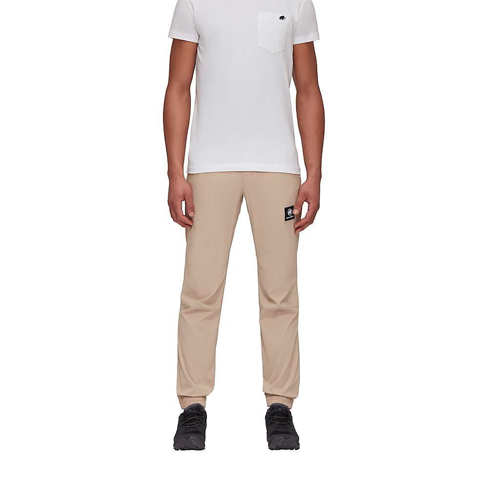 A person is wearing a white short-sleeve t-shirt, Mammut Men's Massone Light Pant in eco-friendly beige with a black logo patch on the left leg, and black shoes, standing against a plain white background.