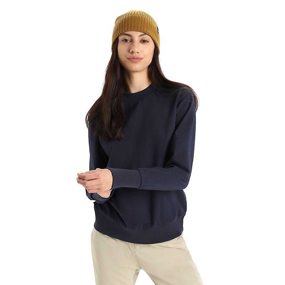 A woman with long brown hair and a neutral expression stands against a plain white background wearing the Icebreaker Womens Central II LS Sweatshirt in dark navy, a mustard yellow knit beanie, and beige pants.