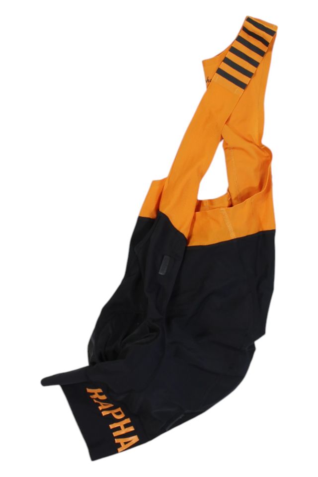 The Rapha Mens Pro Team Bib Shorts II - Regular are orange and black with branded legs, orange shoulder straps featuring black stripes, a race-grade chamois, and are displayed flat on a white background.