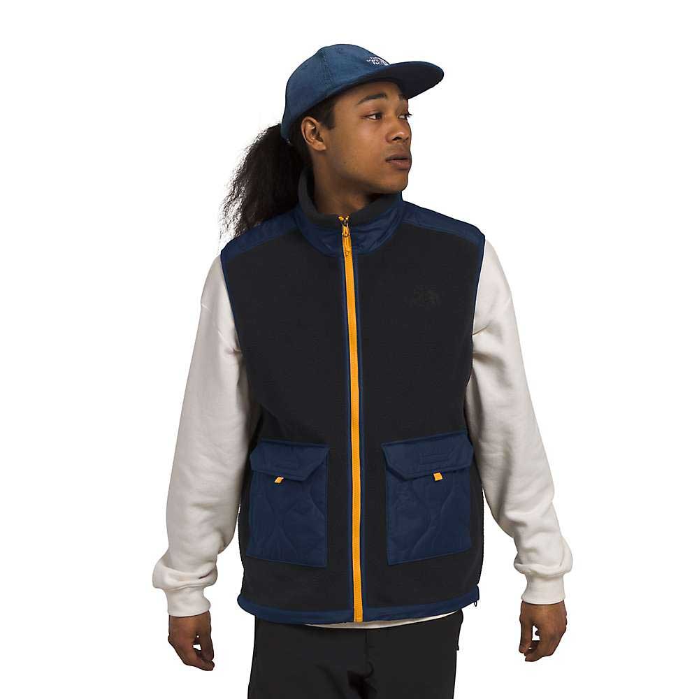 A person in a blue cap and white long-sleeve shirt wears The North Face Men's Royal Arch Vest, navy with yellow zipper and pockets, made from recycled polyester, standing against a white background looking to the side.