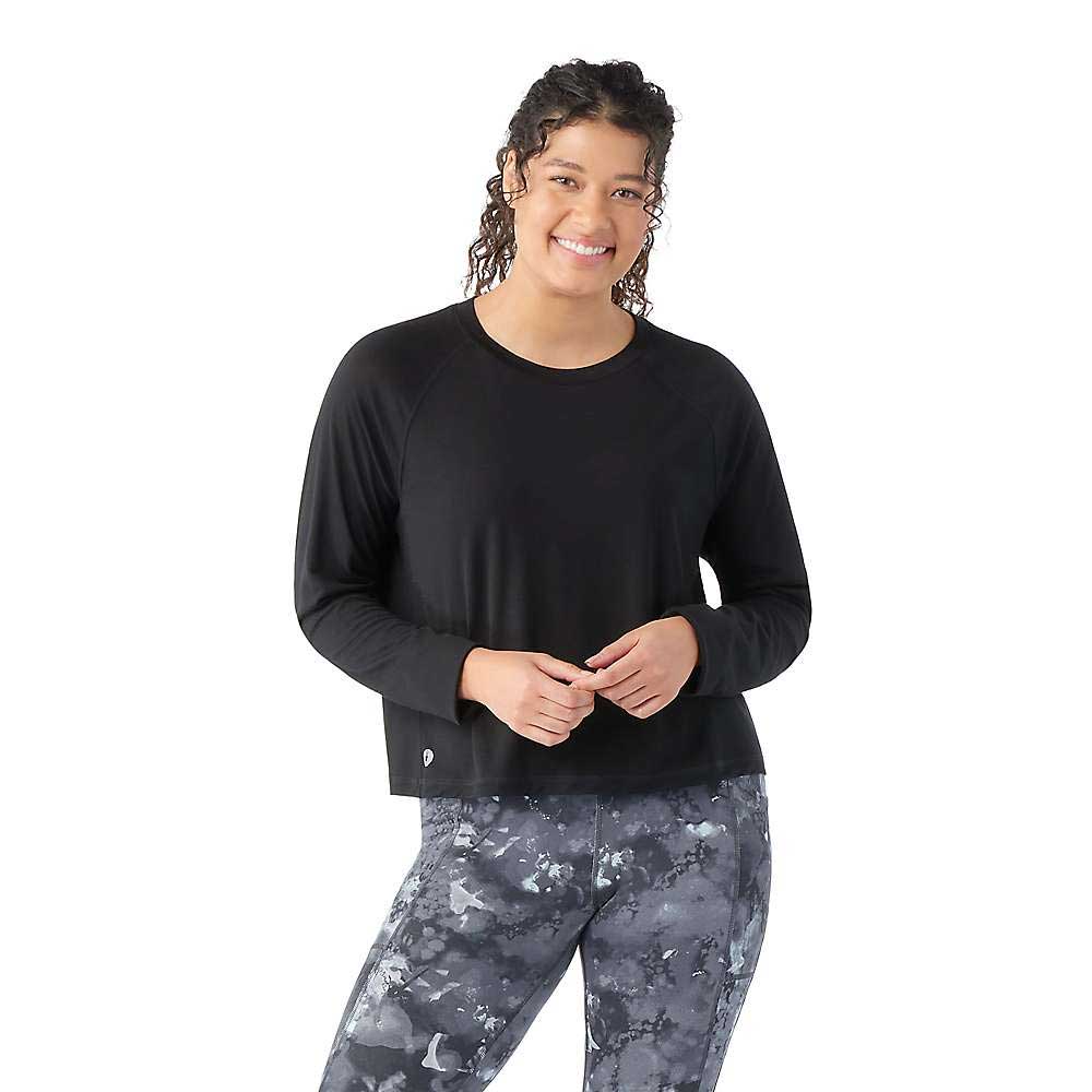 A woman with curly hair smiles while wearing the Smartwool Women's Active Crop LS Top, made from lightweight performance fabric, and stands against a plain white background.