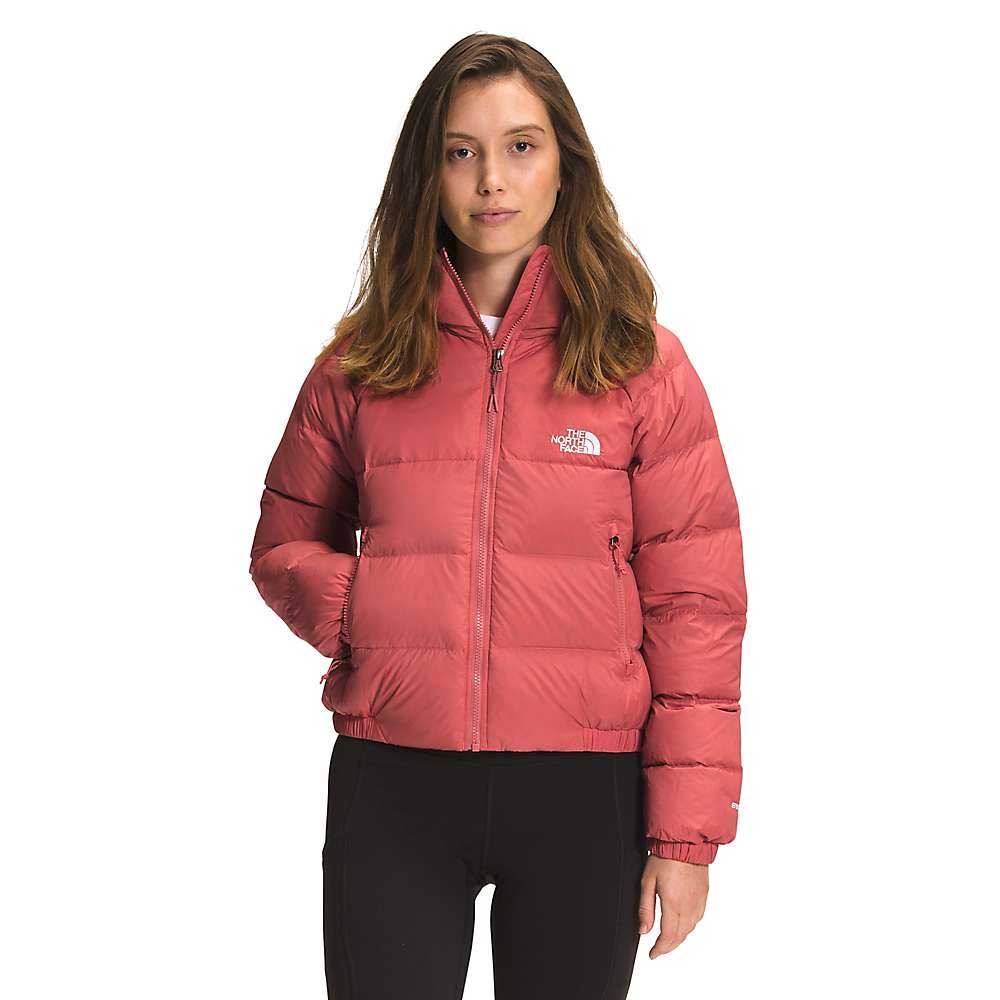 A woman wears The North Face Womens Hydrenalite Down Hoodie in pink, featuring the logo, and stands with one hand in her pocket against a plain white background.