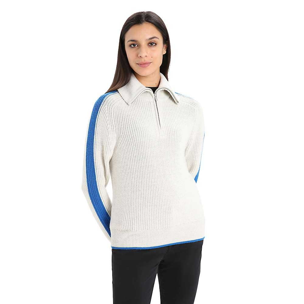 A woman stands facing the camera against a white background, wearing the Icebreaker Womens Lodge LS Half Zip Sweater in cream with blue sleeve stripes, her hands behind her back.