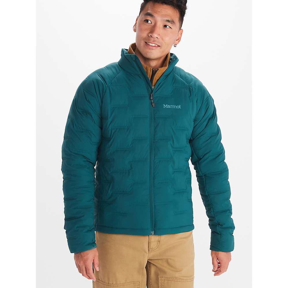 A man wears a teal Marmot Mens WarmCube Active Novus Jacket and tan pants, standing forward with a slight smile, looking off to the side against a plain white background.