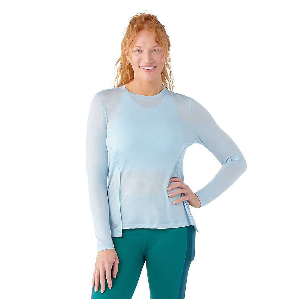 A woman with long red hair wears a light blue Smartwool Womens Active LS Crew Top and dark teal leggings, standing with one hand on her hip and smiling against a white background.