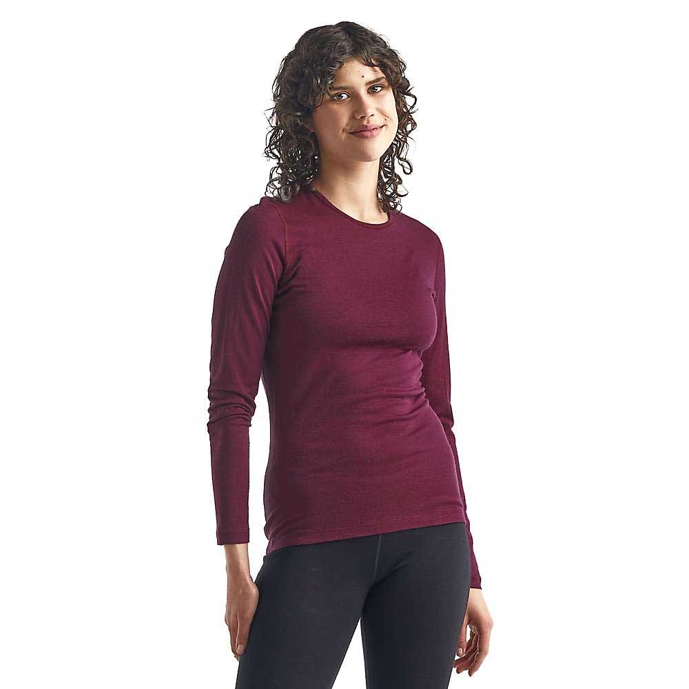 A woman with curly hair wears the Icebreaker Womens 200 Oasis LS Crewe and black pants, standing against a plain white background and smiling slightly.