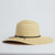 The Coal Seaside Hat is a wide-brimmed, light tan straw sun hat with a rounded crown and thin, dark bow-tied band—perfectly packable for sunny adventures.