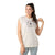 A woman with long dark hair in a ponytail smiles against a white background while wearing the Smartwool Womens Active Muscle Tank, a sleeveless off-white top featuring a colorful sun graphic and "Go Far. Feel Good." slogan, made from moisture-wicking Merino wool.