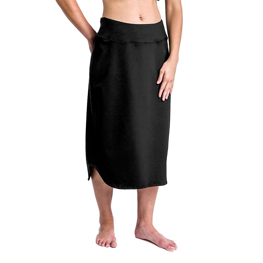 A person stands barefoot wearing the Stonewear Designs Womens Cirrus Skirt in black, a knee-length skirt with a wide waistband and slight side slit. Hands rest on the skirt; upper body is partially out of frame.