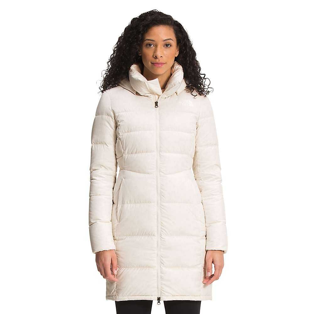 A woman with curly hair wears The North Face Womens Metropolis Parka, a long, hooded down jacket in a light color, standing facing forward against a plain white background.