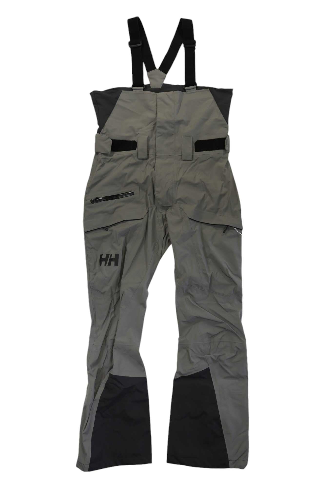 The Helly Hansen Men's Sogn Bib Shell Pant in gray and black features suspenders, multiple pockets, adjustable straps, and reinforced knees and ankles for skiing. A small HH logo is on the left thigh.