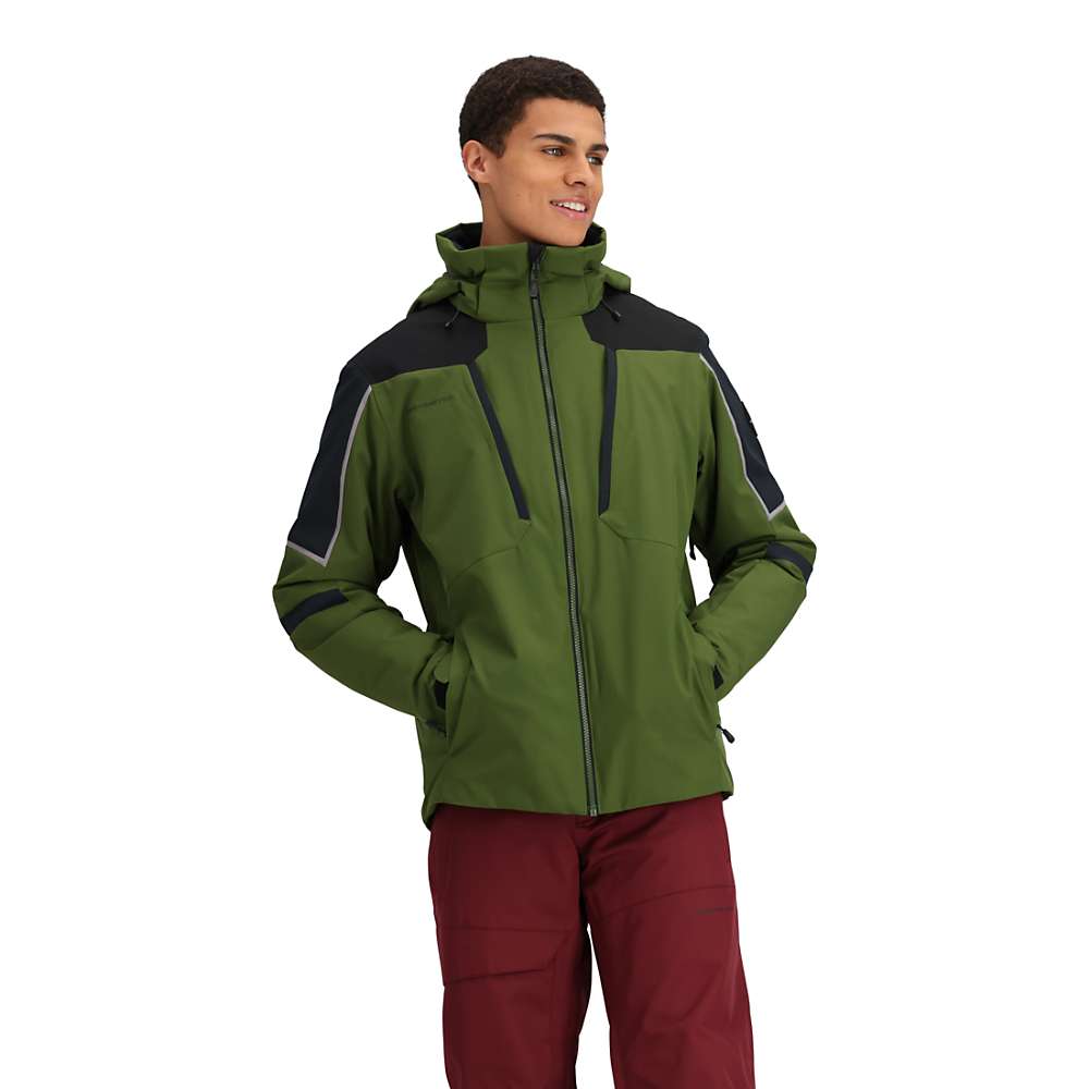 A person wears the Obermeyer Mens Foundation Jacket in green with black accents and red pants, standing with hands in pockets, smiling and looking to the side against a plain white background.