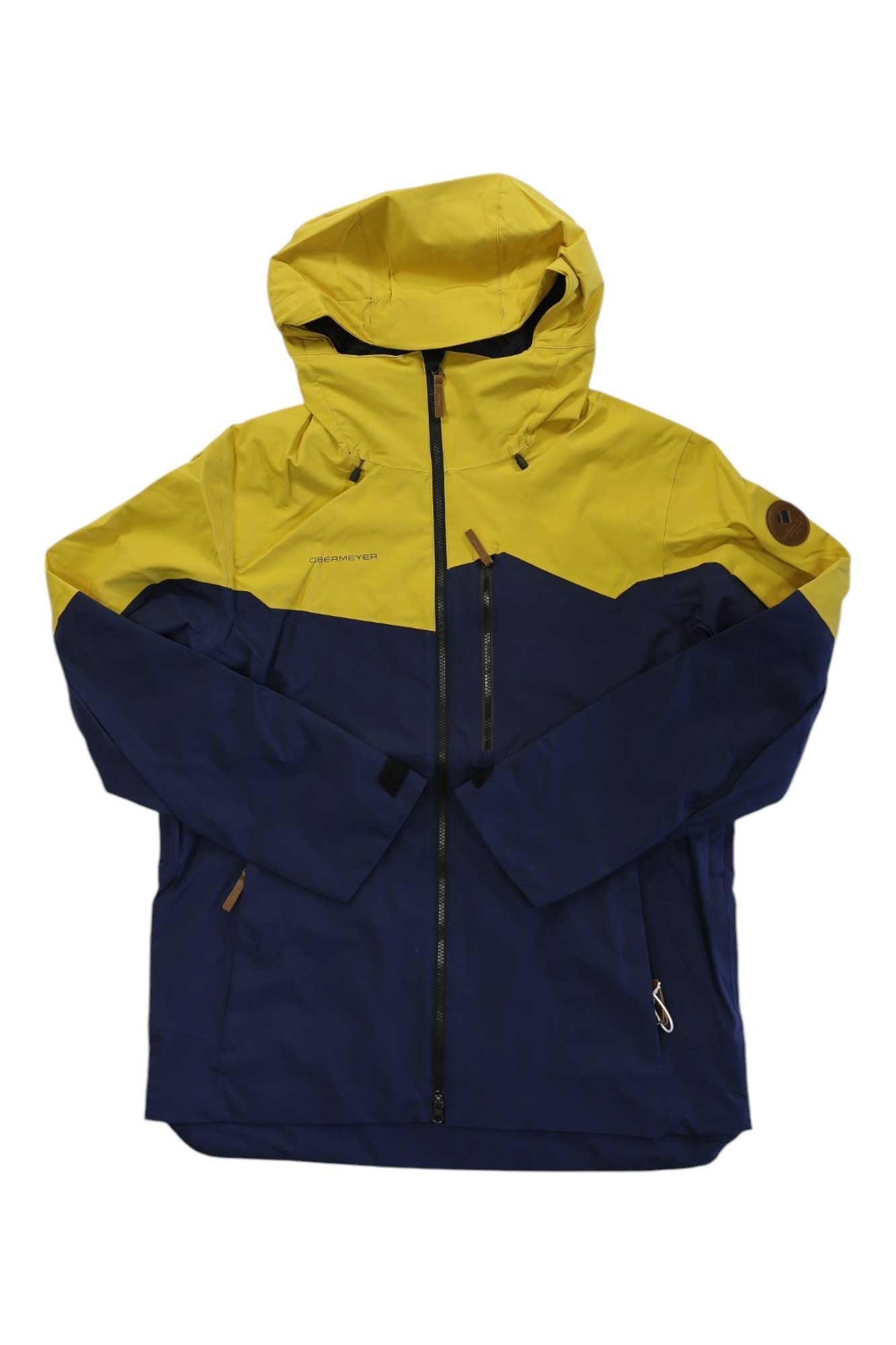 The Obermeyer Mens Chandler Shell Jacket is a water-resistant outdoor shell with a yellow top and sleeves, navy blue bottom, front zipper, zippered pockets, and a small round brown logo patch on the left sleeve. Perfect for mountain adventures.