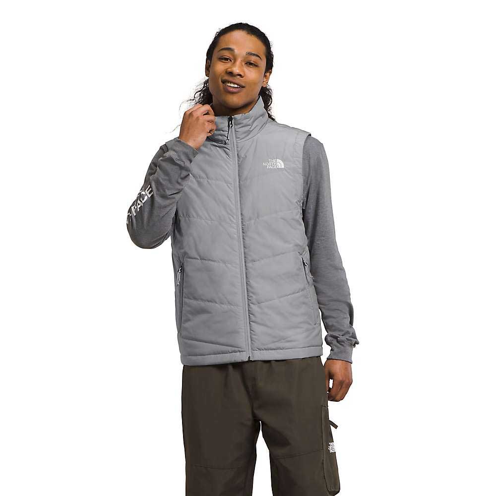 A person smiles, touching their neck, wearing a light gray The North Face Men's Junction Insulated Vest with recycled insulation over a gray long-sleeve shirt and dark olive cargo pants. Both the vest and shirt display The North Face logo.