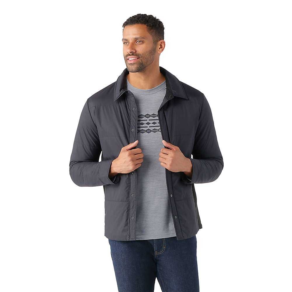 A man wears a dark gray Smartwool Mens Smartloft Shirt Jacket over a light gray graphic tee and blue jeans, facing slightly right, holding his jacket edges and smiling against a white background.