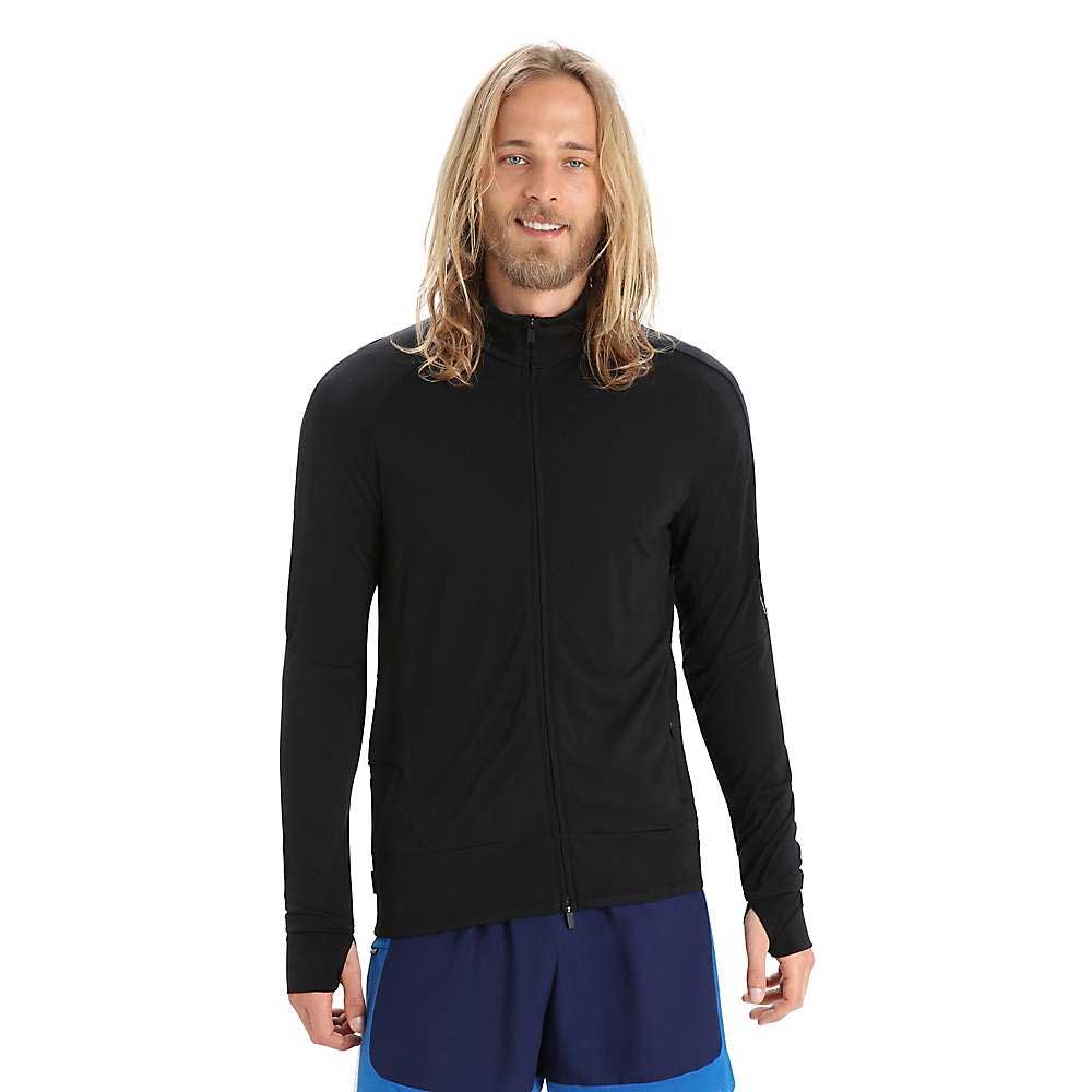 A smiling person with long blond hair wears the Icebreaker Mens ZoneKnit LS Zip Jacket in black and blue shorts, standing against a plain white background.