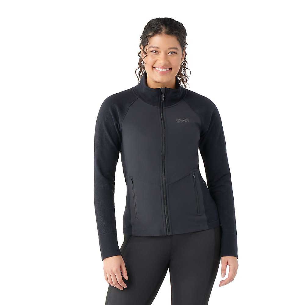 A woman with long curly hair smiles against a plain white background, wearing the Smartwool Womens Intraknit Active Full Zip Jacket and black pants.