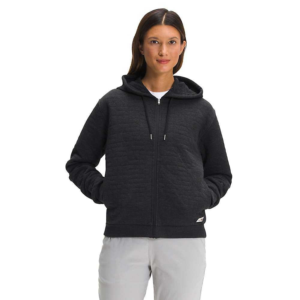 A woman faces forward with her hands in the pockets, wearing The North Face Womens Longs Peak Quilted Full Zip Hoodie in black and light gray pants, set against a plain white background.