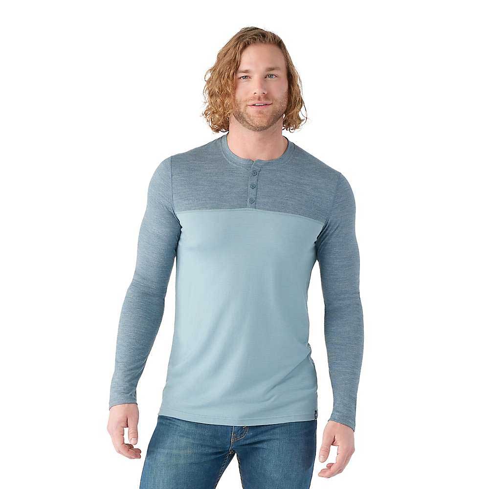 A man with wavy, shoulder-length blond hair wears the Smartwool Mens Merino Sport 150 Henley, featuring moisture-wicking fabric and a textured blue yoke and sleeves, paired with blue jeans, standing against a white background.