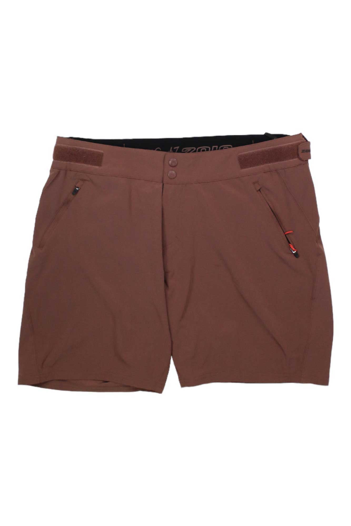 The Zoic Womens Navaeh 7 Short is a brown cycling short featuring two front zippered pockets, button and zip closure, adjustable waist tabs, and a packable design for easy travel.