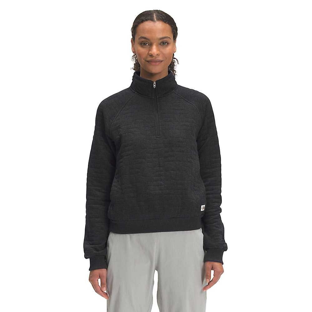 A woman with braided hair, smiling against a white background, wears The North Face Women's Longs Peak Quilted 1/4 Zip Jacket in black and light gray pants.