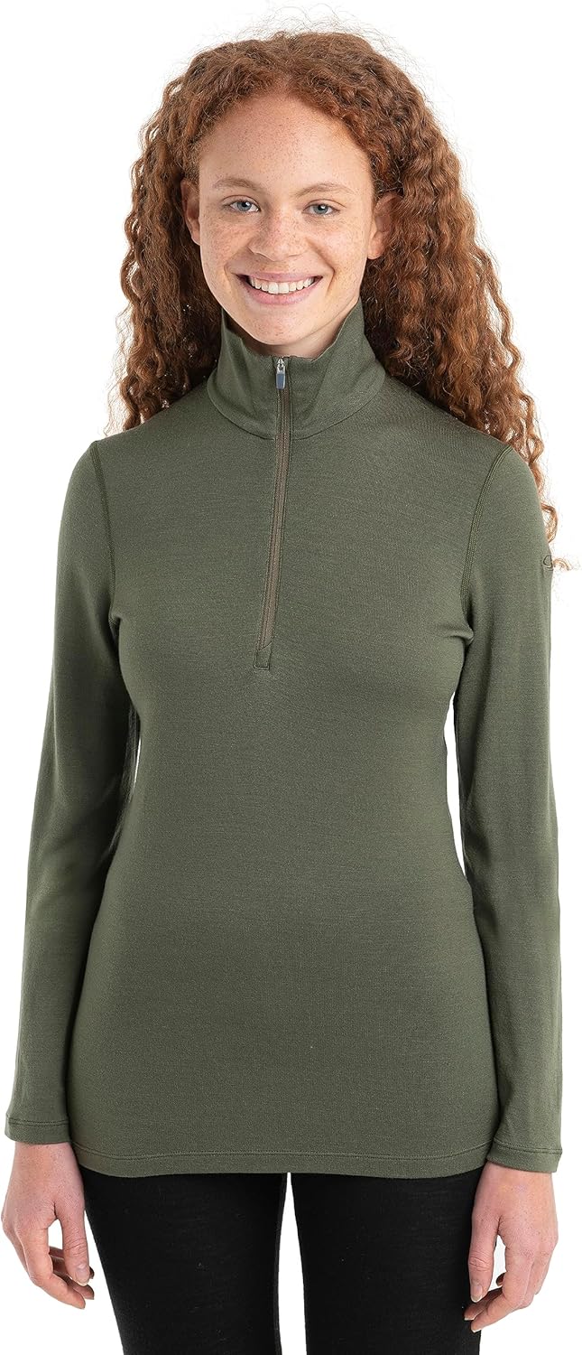 A woman with long curly red hair smiles, wearing the Icebreaker Womens 260 Tech LS Half Zip Top in olive green and black pants against a white background—an effortlessly stylish choice for outdoor wear.