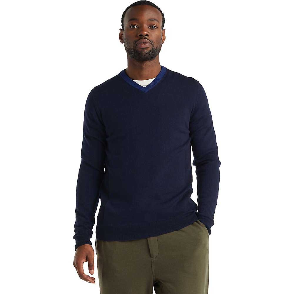 A man faces forward against a plain white background, wearing the Icebreaker Mens Shearer V Sweater in navy blue over a blue undershirt and olive green pants, with one hand in his pocket.