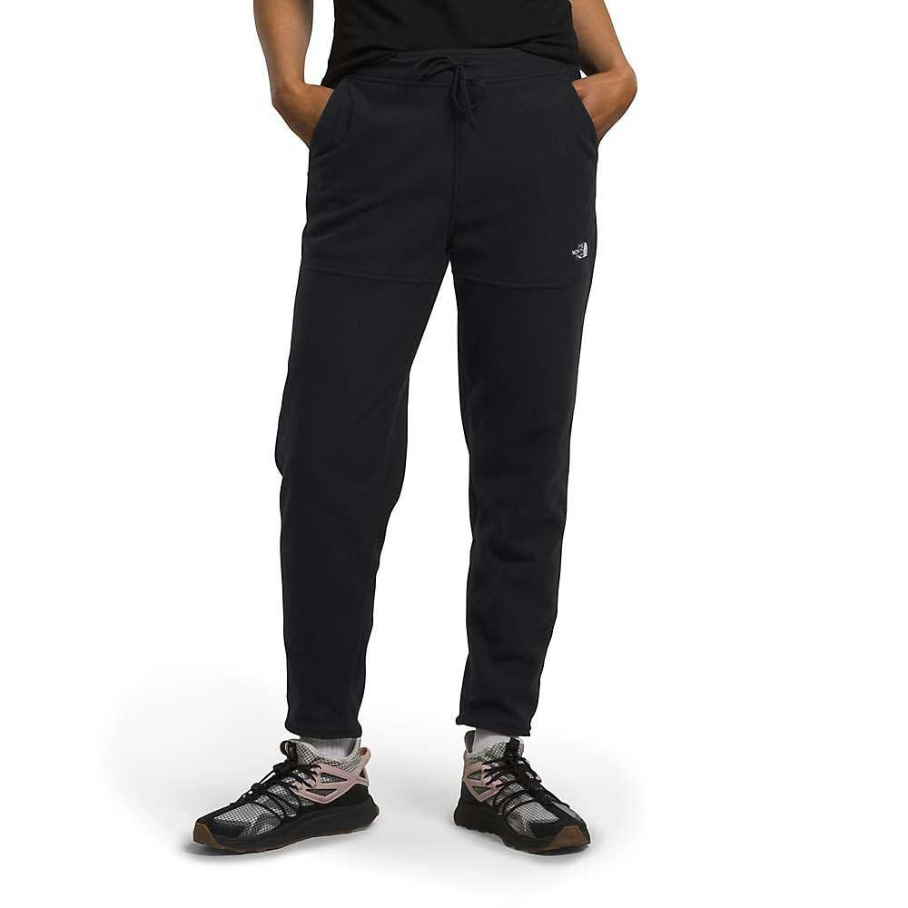 A person stands with hands in pockets against a white background, wearing The North Face Womens Alpine Polartec 100 Pant in black, a black t-shirt, gray socks, and black athletic shoes with pink laces.
