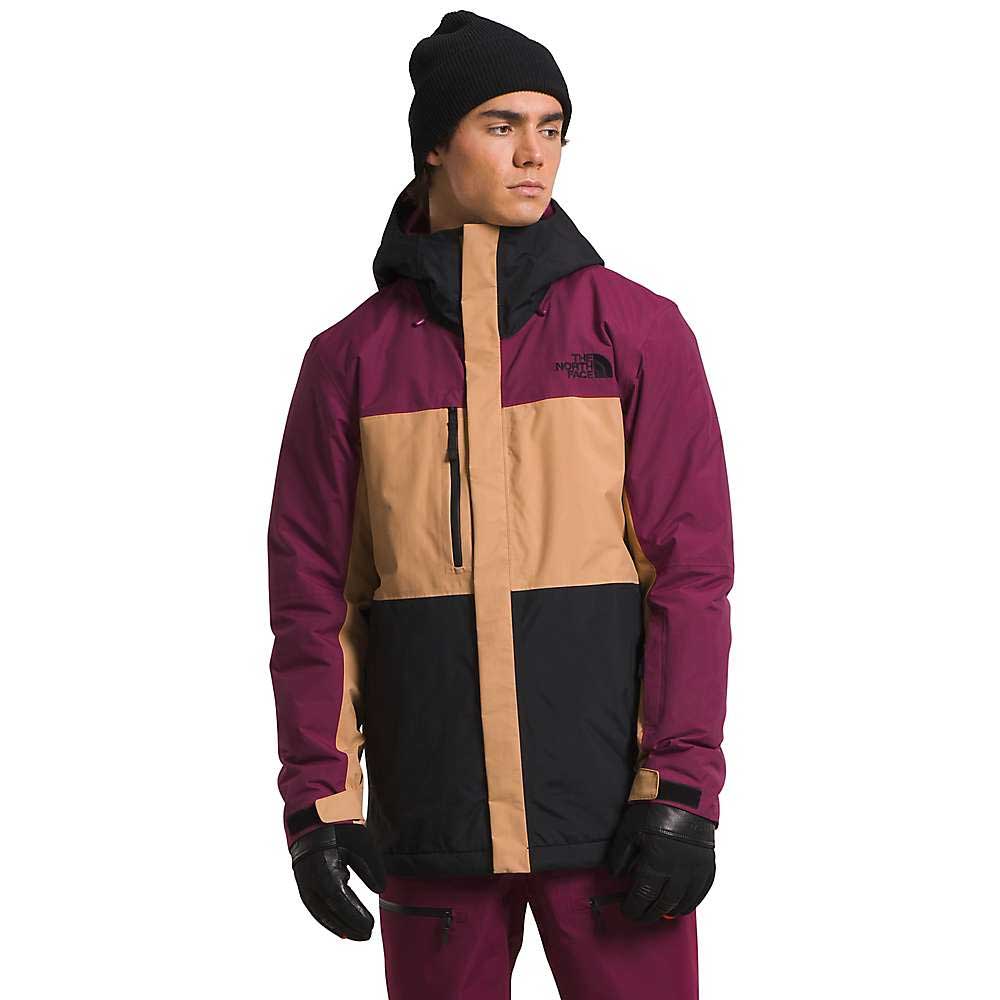 Wearing The North Face Men's Freedom Insulated Jacket in maroon, tan, and black—with Heatseeker Eco insulation—a black beanie, maroon pants, and black gloves, a person stands against a white background with the hood up, looking to the side.