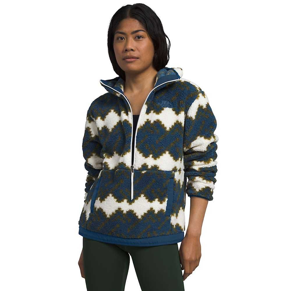 A person wears The North Face Womens Campshire Fleece Hoodie in blue, white, and olive green zigzag pattern, paired with dark green pants and a white undershirt, standing with hands at their sides and looking forward.