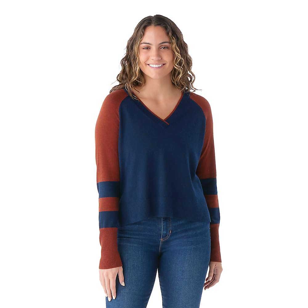 A woman with wavy hair smiles while wearing a Smartwool Womens Edgewood V-Neck Sweater with rust sleeves and navy stripes, paired with blue jeans, standing in front of a white background.