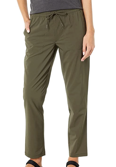 Lower body of a woman shown wearing The North Face Womens Never Stop Wearing Ankle Pant in olive green recycled polyester, styled with a dark top and black sandals, one hand in the pocket—ideal activewear against a plain background.