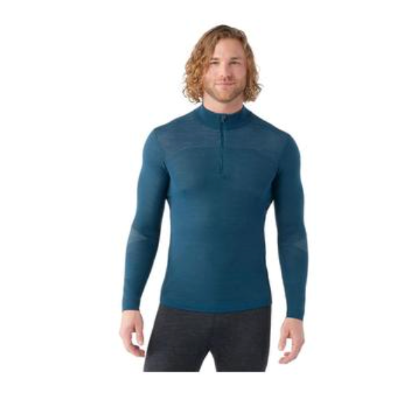 A man with wavy, shoulder-length blond hair is wearing a fitted Smartwool Men's Intraknit Merino 200 1/4 Zip Top and dark pants, standing and smiling against a plain white background.