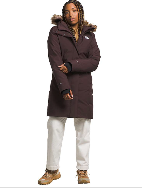 A person stands facing forward, one hand by their side and the other partly tucked in their sleeve, wearing The North Face Womens Arctic Parka—long, dark brown with a fur-lined hood—paired with light pants and tan boots.