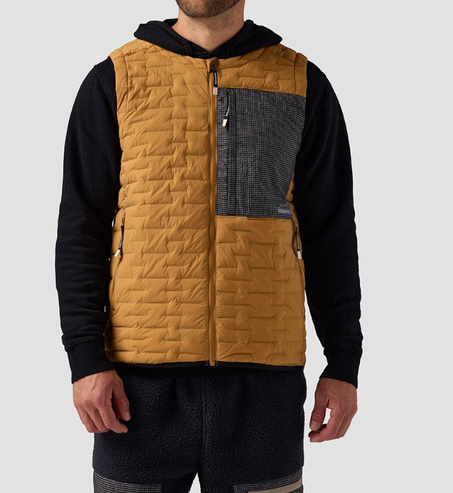 A man wears the Backcountry Peale Primaloft Stitch-Free Baffle Stretch Vest—featuring lightweight insulation and a black patterned chest pocket—over a black hoodie and dark textured pants, standing against a light background.